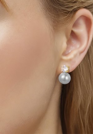 Primer plano de un pendiente de oro con una piedra preciosa clara y una perla plateada grande llevado en una oreja de piel clara con cabello castaño claro.