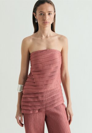 Mujer con top rosa empolvado sin tirantes y en capas, pantalones a juego, brazalete de plata en la parte superior del brazo y pendientes de gota sobre fondo liso.