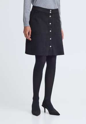 Kobieta w czarnej spódnicy o linii A z guzikami z przodu, czarnych rajstopach, szarym długim rękawie i czarnych szpilkach na białym tle.