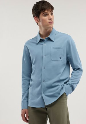 Giovane uomo che indossa una camicia celeste a maniche lunghe con taschino sul petto, in piedi con una mano nella tasca dei pantaloni verde oliva.