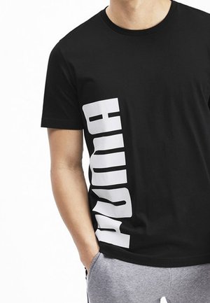 Man wearing black Puma T-shirt with large white vertical logo on the left side and gray sweatpants, standing with one hand in pocket.