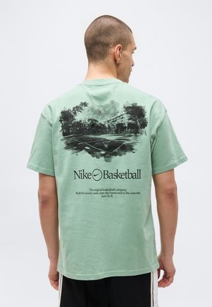 Hombre con pelo corto que lleva una camiseta verde de Nike Basketball que muestra un gráfico en blanco y negro de una cancha de baloncesto y texto en la espalda.