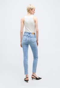 Άτομο με κοντά ξανθά μαλλιά που φοράει λευκό αμάνικο τοπ, ανοιχτό μπλε τζιν με ψηλή μέση και μαύρα τακούνια slingback, γυρισμένο πλάτη.