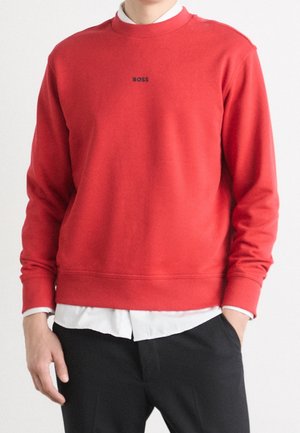 Person trägt ein rotes Langarm-Sweatshirt mit dem "BOSS"-Logo, darunter ein weißes Hemd und schwarze Hosen, die Hände teilweise in den Taschen.