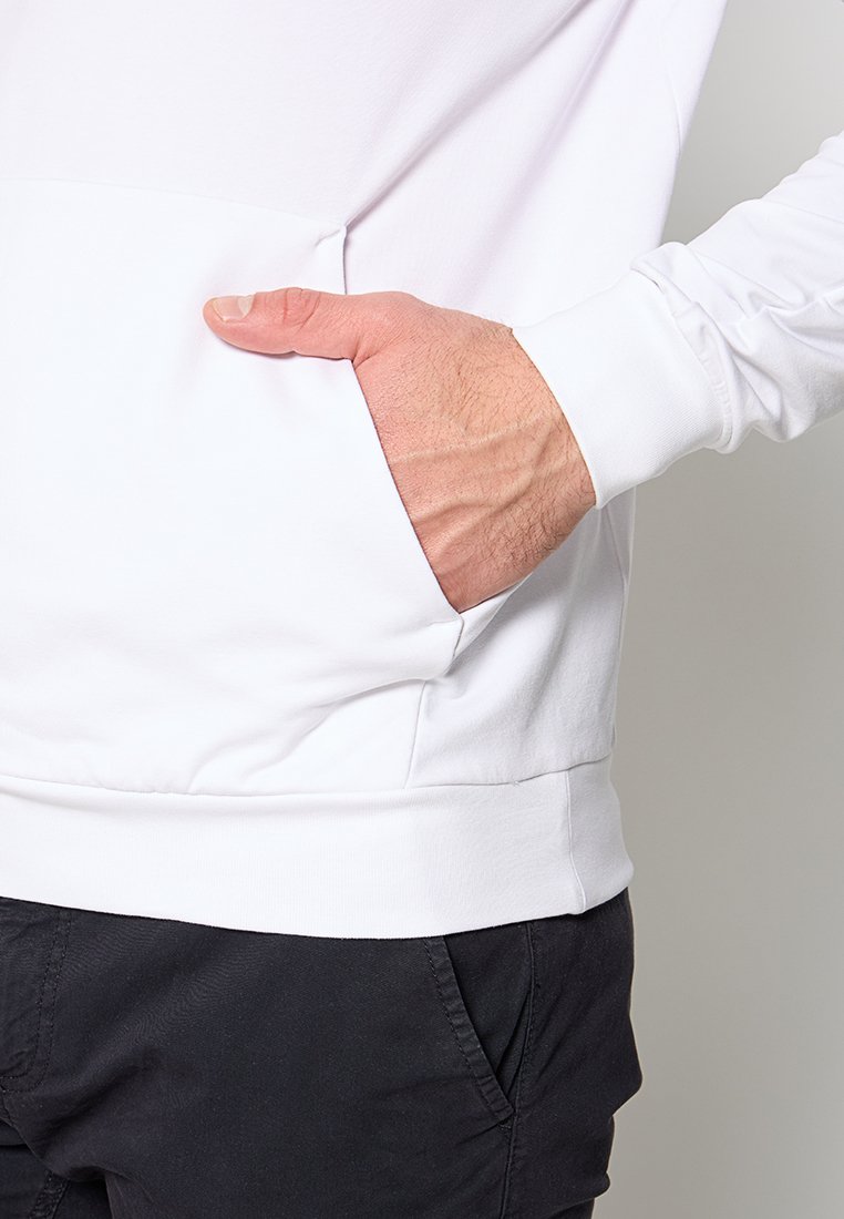 Hombre vistiendo un sudadera blanca con la mano descansando dentro del bolsillo delantero, combinada con pantalones negros.