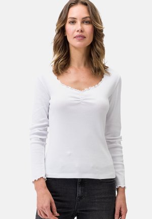 Weißes Langarmshirt mit Rundhalsausschnitt und Rüschensaum, aus geripptem Stoff, kombiniert mit dunklen Jeans, die an der Vorderseite eine dezente Raffung aufweisen.