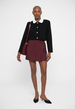 Veste noire courte avec col blanc, boutons argentés, associée à une mini-jupe bordeaux. Le modèle tient un sac bordeaux et porte des mules noires.
