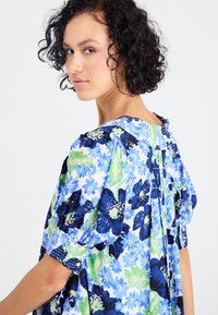 Bloemenjurk met blauwe en groene bloemen op een lichte achtergrond. Pofmouwen en een knoopsluiting aan de achterkant verbeteren het ontwerp.