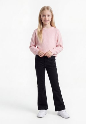 Ung pige med langt blondt hår iført en lyserød trøje, sorte bukser og hvide sneakers, der står og smiler mod en hvid baggrund.