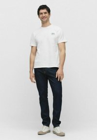 Hvid bomulds t-shirt med et lille grønt print, kombineret med mørkeblå denim jeans og beige sneakers. Enkel, afslappet pasform.