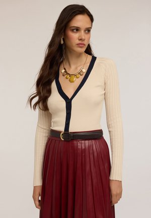 Donna che indossa un cardigan a coste color crema con scollatura a V e rifiniture nere, una collana voluminosa, e una gonna plissettata in pelle bordeaux con una cintura nera.
