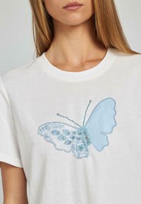T-shirt bianco in cotone con un grande design di farfalla blu ricamata, arricchita da perline dettagliate ed elementi decorativi sulle sue ali.