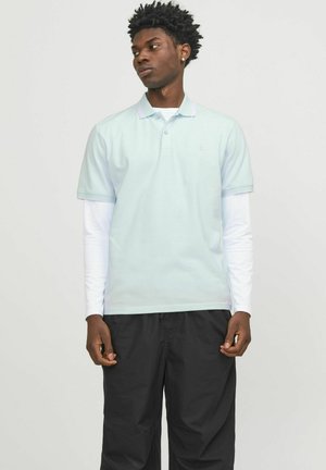 Hellsblaues Polohemd mit weicher Textur, mit Kragen und kurzen Ärmeln, über einem weißen Langarmshirt getragen. Schwarze Hose vervollständigt das Outfit.