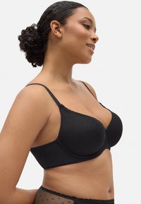 Sujetador negro acolchado con acentos de encaje, textura de lunares, tirantes ajustables y un diseño sin costuras, mostrado de lado sobre un fondo neutro.