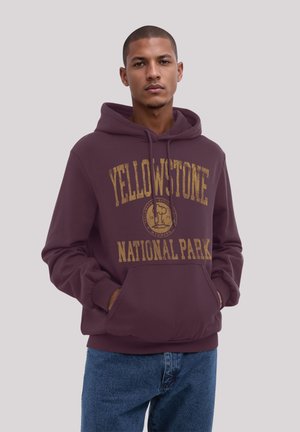 Burgundy hoodie with a drawstring hood, featuring "YELLOWSTONE NATIONAL PARK" in distressed gold print and a front pocket. Denim jeans visible.
