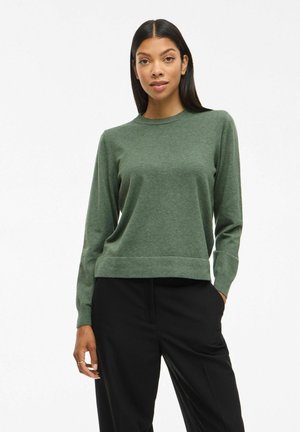 Pull en tricot vert avec un col rond et des manches longues, doté d'un ourlet et de poignets côtelés. Texture lisse ; associé à un pantalon noir.