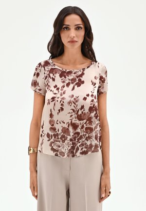 Femme aux longs cheveux foncés portant un haut beige à manches courtes avec un imprimé floral marron et un pantalon beige clair à jambes larges sur un fond uni.