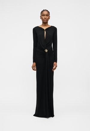 Γυναίκα που φοράει μακρύ μαύρο φόρεμα με μακριά μανίκια, λαιμόκοψη με άνοιγμα και χρυσές λεπτομέρειες στο στήθος και τη μέση, στέκεται μπροστά σε απλό λευκό φόντο.