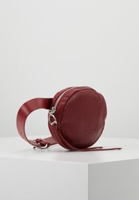 Borsa rotonda in pelle rossa con superficie texturizzata, chiusura con zip e tracolla regolabile. Presenta un design elegante con hardware minimale.