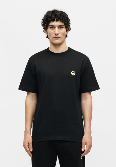 Άνδρας με σγουρά μαλλιά που φοράει μαύρο μπλουζάκι και παντελόνι με μικρό κίτρινο έμβλημα θλιμμένου προσώπου στο στήθος και στον μηρό.