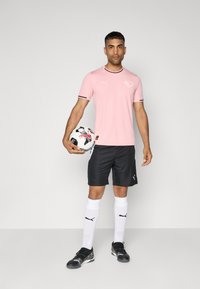 T-shirt rose clair avec des accents noirs, shorts noirs, chaussettes blanches et chaussures noires. Tenant un ballon de football noir et blanc, debout sur un fond neutre.