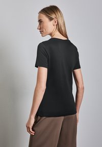 Sort kortærmet T-shirt med rund halsudskæring, glat tekstur og tætsiddende design. Båret med brune bukser, set fra ryggen.