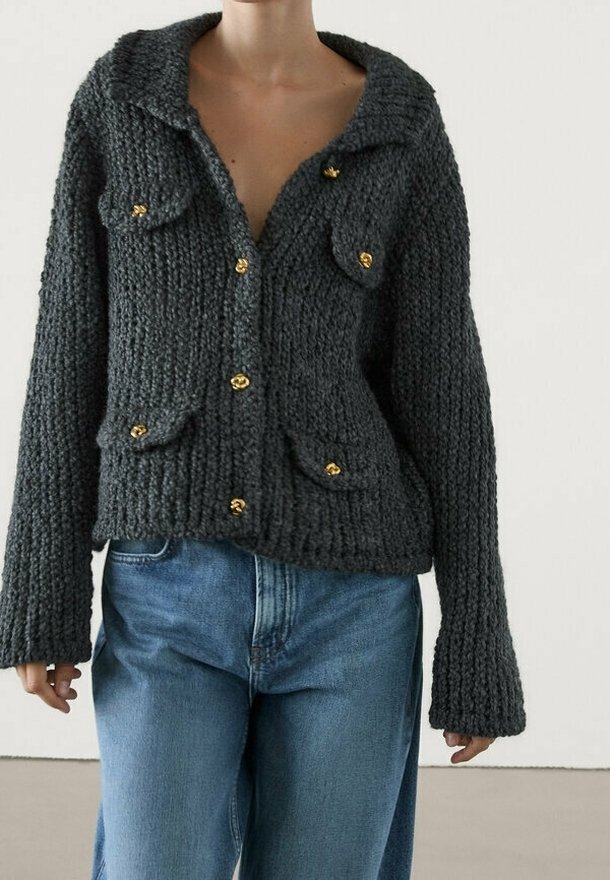 Cardigan en tricot épais gris foncé avec boutons dorés et quatre poches à rabat sur le devant, porté avec un jean bleu clair par une personne debout.