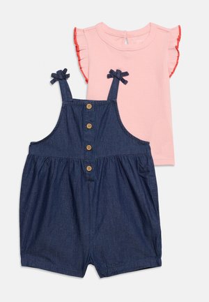 Chemise rose à manches courtes avec bords rouges volantés, associée à une combinaison short en denim bleu marine avec boutons sur le devant et bretelles nouées aux épaules.