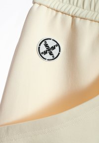 Tejido beige claro con una textura suave, presenta un parche bordado circular en blanco y negro. Detalle de cinturilla elástica.