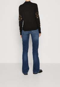 Pull noir avec broderie décorative d'un flocon de neige doré sur les manches, associé à un jean bleu évasé et des chaussures noires. Vue de dos affichée.