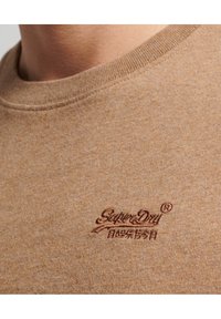 Närbild av en ljusbrun crewneck-tröja med en liten broderad "SuperDry"-logotyp och japanska tecken på bröstet.