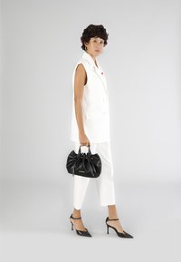 Valkoinen ilmanhihoinen bleiseri yhdistettynä valkoisiin housuihin, mustaan käsilaukkuun ja mustiin kärkitossuihin. Sileä pinta minimalistisella suunnittelulla.