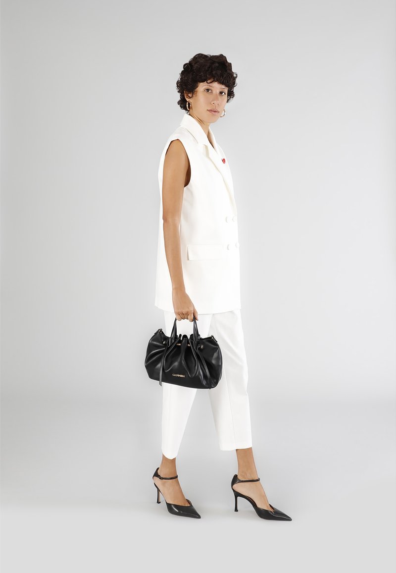 Valkoinen ilmanhihoinen bleiseri yhdistettynä valkoisiin housuihin, mustaan käsilaukkuun ja mustiin kärkitossuihin. Sileä pinta minimalistisella suunnittelulla.