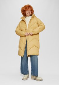 Geel pufferjack met chevron-quilting, frontale rits, verstelbare capuchon en twee frontzakken. Draagt over een crèmekleurige trui en blauwe jeans.