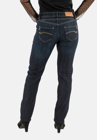 Jeans de denim azul oscuro con corte ajustado, diseño de bolsillos traseros único con costuras y remaches dorados, terminados con una cinturilla estándar.