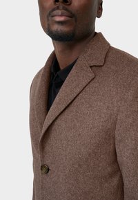 Bruine wolmix jas met een geknipte kraag, enkele knoopsluiting en een textuurstof. Draagt over een zwart shirt.