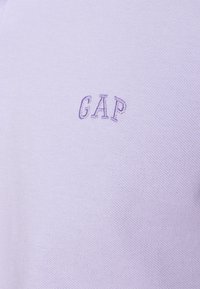 Pull court à manches courtes en tricot violet clair avec petit logo "GAP" brodé en violet plus foncé sur la poitrine. Tissu texturé.