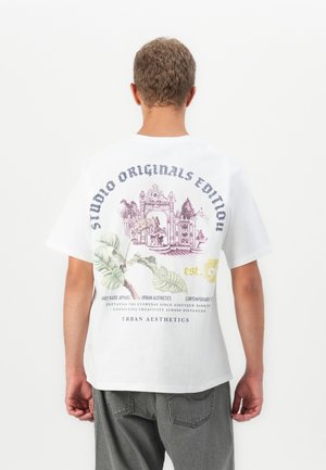 Ung person med kort, krøllet hår iført en hvid T-shirt med et blomster- og arkitektonisk design samt tekst på ryggen, kombineret med grå jeans.