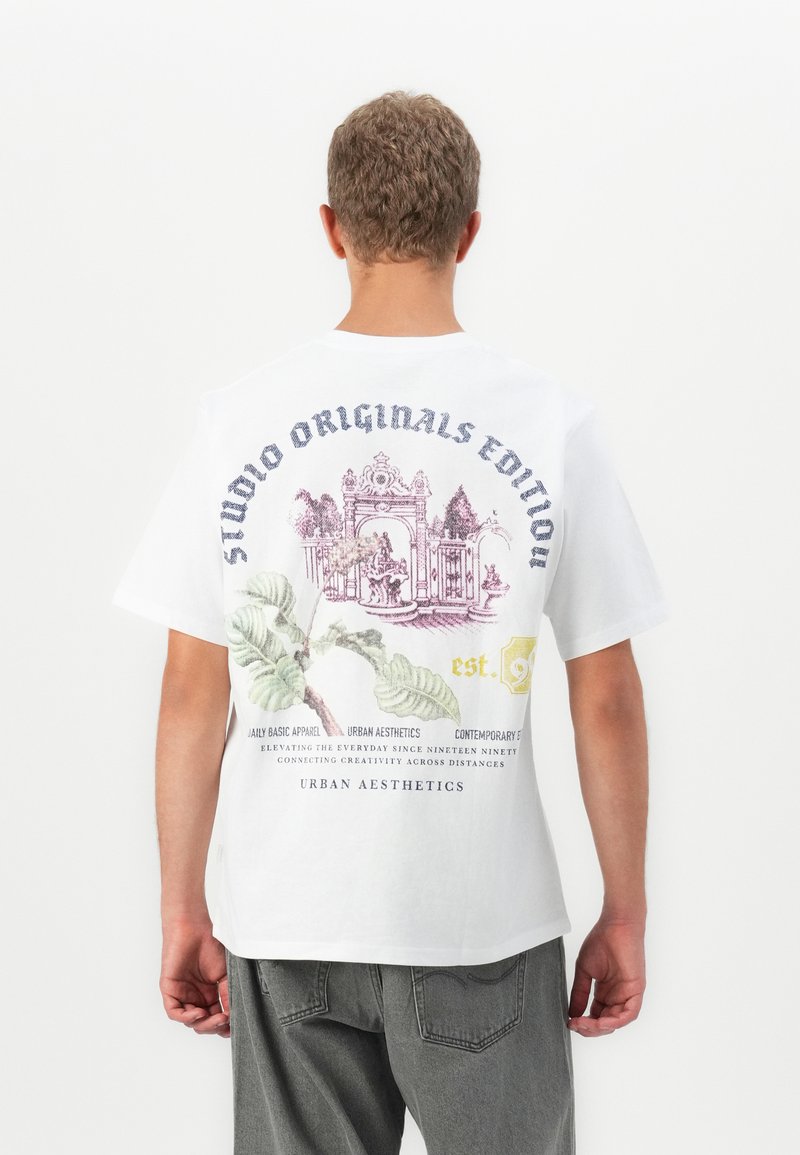 Ungdom med kort krøllete hår som har på seg en hvit T-skjorte med et blomster- og arkitektonisk design med tekst på ryggen, sammen med grå jeans.