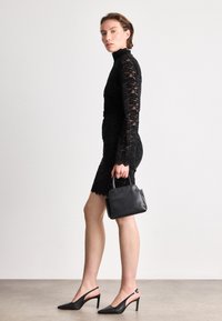 Μαύρο φόρεμα με δαντέλα και μακριά μανίκια, με εφαρμοστό σιλουέτα και κομψό τελείωμα, συνδυασμένο με μαύρες δερμάτινες γόβες με slingback και μια μικρή μαύρη τσάντα.