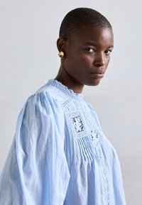 Vaaleansininen puuvillapaita, jossa on laskostetut hihat, pitsiyksityiskohtia ja brodeerauksia. Mallilla on päässään pyöreät kultaketjut.