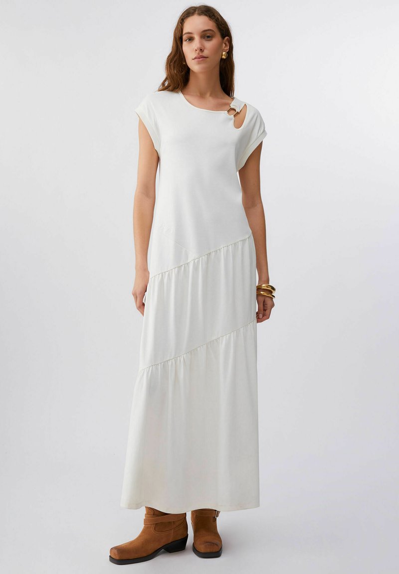 Robe longue blanche à manches courtes, avec un détail découpé à l'épaule et un ourlet à volants superposés. Le mannequin porte des bottines marron.