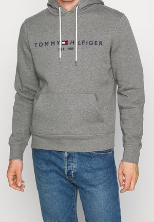 Mężczyzna w szarej bluzie Tommy Hilfiger z logo i niebieskich dżinsach, stojący z rękami swobodnie opuszczonymi wzdłuż ciała.