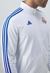 Hvit idrettsjakke med blå Adidas-striper på skuldrene, glidelås og Real Madrid-logo på brystet. Glatt stoffstruktur.