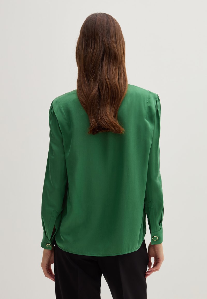 Frau mit langen braunen Haaren, die eine grüne Bluse mit langen Ärmeln und schwarze Hosen trägt, steht mit dem Rücken zur Kamera vor einem weißen Hintergrund.