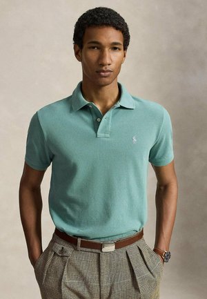 Jeune homme aux cheveux courts et bouclés portant un polo vert clair et un pantalon marron à carreaux, les mains dans les poches.