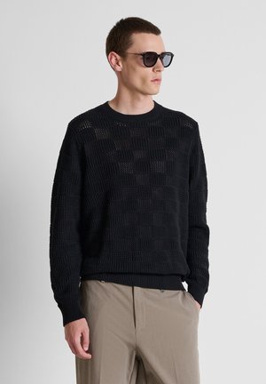 Mann trägt schwarzen texturierten Strickpullover, beige Hose und dunkle Sonnenbrille und schaut seitlich vor einem einfarbigen grauen Hintergrund.