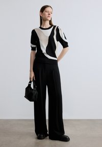 Pletený top s abstraktnými čiernymi, béžovými a bielymi vzormi; doplnený o čierne široké nohavice a malú čiernu textúrovanú kabelku.