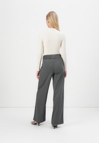 Femme portant un haut blanc à manches longues et un pantalon gris ample taille haute, debout de dos devant un fond blanc.