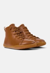 Botas de tobillo de cuero marrón con una textura suave, puntera redondeada y sistema de cordones; forro suave visible en la parte superior y suela flexible.